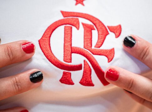 Close no escudo do Flamengo em uma camisa branca de jogo. Perto do escudo estão dedos com unhas pintadas de preto ou vermelho