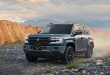 Um SUV robusto percorre uma estrada de terra em uma paisagem montanhosa. O veículo é moderno, com design quadrado e imponente, faróis retangulares e pintura cinza. Ao fundo, surgem montanhas rochosas sob um céu parcialmente nublado.