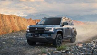 Um SUV robusto percorre uma estrada de terra em uma paisagem montanhosa. O veículo é moderno, com design quadrado e imponente, faróis retangulares e pintura cinza. Ao fundo, surgem montanhas rochosas sob um céu parcialmente nublado.