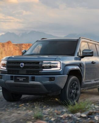 Um SUV robusto percorre uma estrada de terra em uma paisagem montanhosa. O veículo é moderno, com design quadrado e imponente, faróis retangulares e pintura cinza. Ao fundo, surgem montanhas rochosas sob um céu parcialmente nublado.