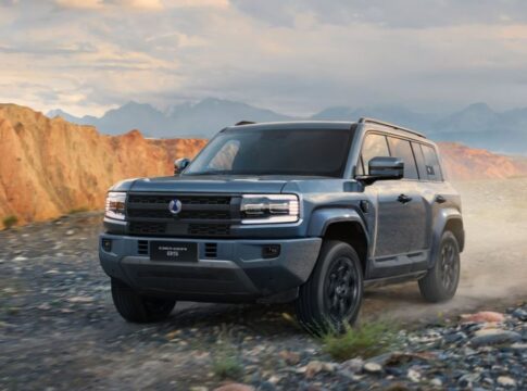 Um SUV robusto percorre uma estrada de terra em uma paisagem montanhosa. O veículo é moderno, com design quadrado e imponente, faróis retangulares e pintura cinza. Ao fundo, surgem montanhas rochosas sob um céu parcialmente nublado.