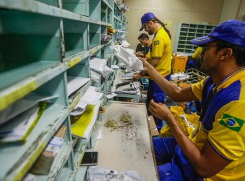 Funcionários dos Correios, uniformizados, manuseiam correspondências dentro de uma sala