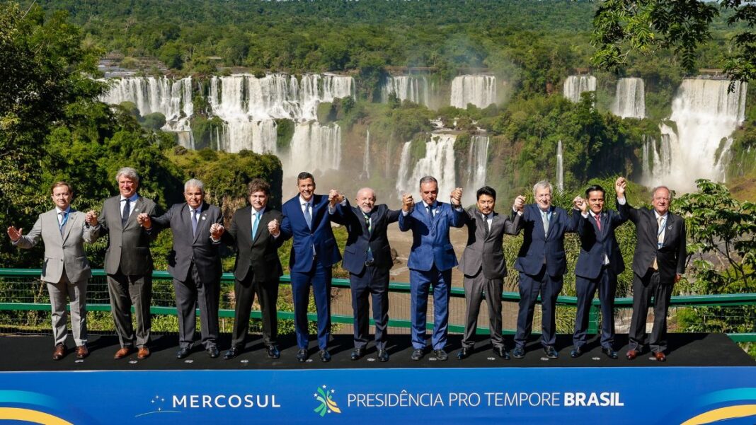 Fotografia oficial dos presidentes e chefes de delegação dos Estados Partes do Mercosul, todos em pé, de mãos dadas, à frente das Cataratas do Iguaçu
