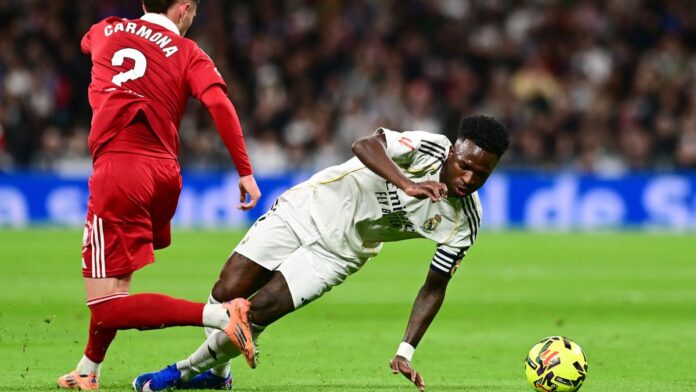 Vini Jr. está caindo no gramado após driblar jogador do Sevilla