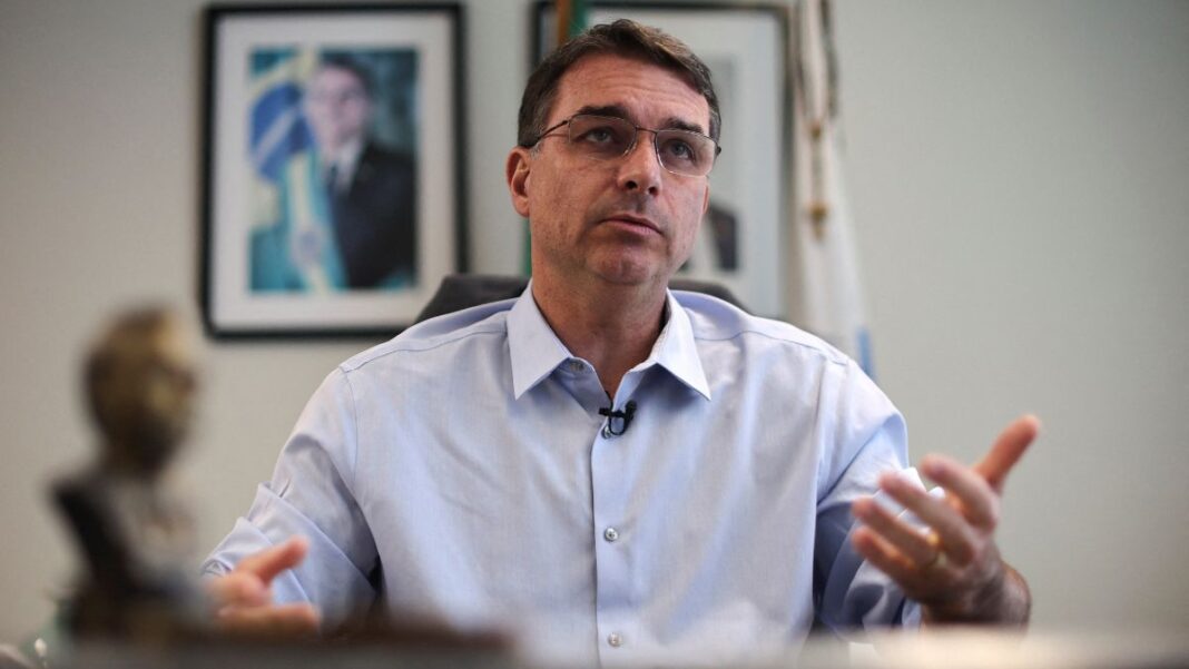 Flávio Bolsonaro gesticula durante entrevista, com foto de Jair Bolsonaro ao fundo