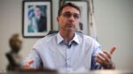 Flávio Bolsonaro gesticula durante entrevista, com foto de Jair Bolsonaro ao fundo