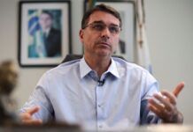 Flávio Bolsonaro gesticula durante entrevista, com foto de Jair Bolsonaro ao fundo