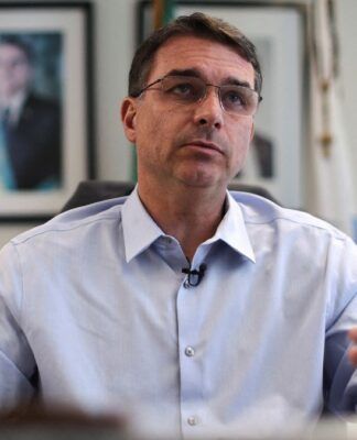 Flávio Bolsonaro gesticula durante entrevista, com foto de Jair Bolsonaro ao fundo