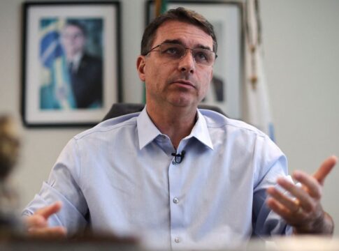 Flávio Bolsonaro gesticula durante entrevista, com foto de Jair Bolsonaro ao fundo