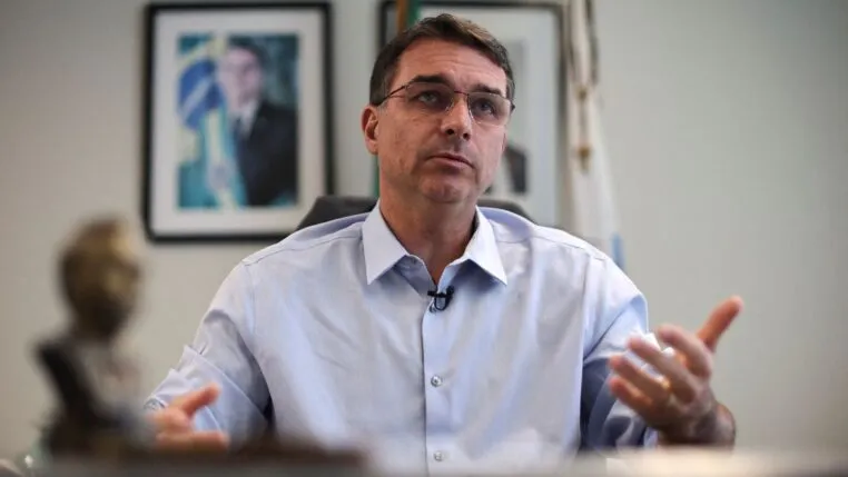 Flávio Bolsonaro gesticula durante entrevista, com foto de Jair Bolsonaro ao fundo
