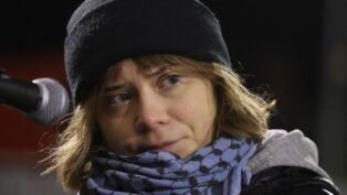 Greta Thumberg veste um gorro preto e um cachecol diante do microfone