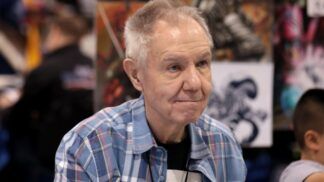 Homem mais velho, de cabelos grisalhos curtos, veste camisa xadrez azul e está sentado em um evento; ao fundo, desfocados, aparecem painéis com ilustrações coloridas e outras pessoas circulando.