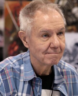 Homem mais velho, de cabelos grisalhos curtos, veste camisa xadrez azul e está sentado em um evento; ao fundo, desfocados, aparecem painéis com ilustrações coloridas e outras pessoas circulando.