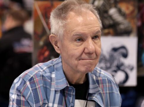 Homem mais velho, de cabelos grisalhos curtos, veste camisa xadrez azul e está sentado em um evento; ao fundo, desfocados, aparecem painéis com ilustrações coloridas e outras pessoas circulando.