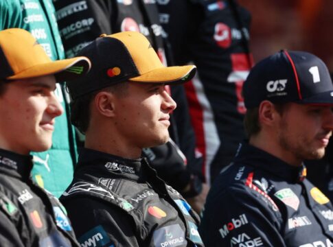 Três pilotos de automóveis sentados lado a lado, usando macacões e bonés de suas equipes de corrida. Eles olham para a frente com expressão neutra, enquanto outros membros de equipes aparecem desfocados ao fundo.