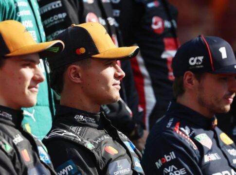 Três pilotos de automóveis sentados lado a lado, usando macacões e bonés de suas equipes de corrida. Eles olham para a frente com expressão neutra, enquanto outros membros de equipes aparecem desfocados ao fundo.