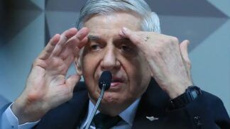 Homem de cabelos grisalhos fala ao microfone em ambiente oficial, gesticulando com as mãos próximas ao rosto enquanto usa terno escuro, camisa branca e gravata verde.