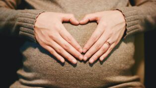 Mãos formando um coração sobre uma barriga de gestante, ambas apoiadas em um suéter de tricô bege; uma das mãos usa um anel delicado.