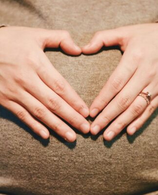 Mãos formando um coração sobre uma barriga de gestante, ambas apoiadas em um suéter de tricô bege; uma das mãos usa um anel delicado.