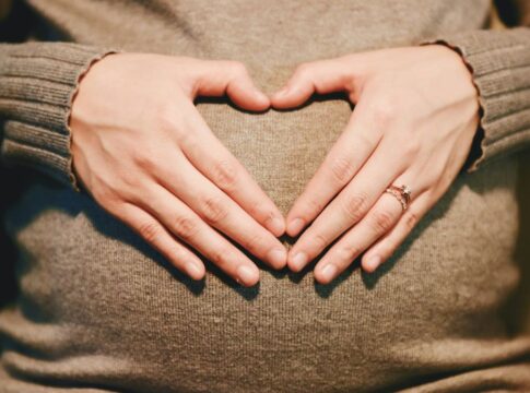 Mãos formando um coração sobre uma barriga de gestante, ambas apoiadas em um suéter de tricô bege; uma das mãos usa um anel delicado.