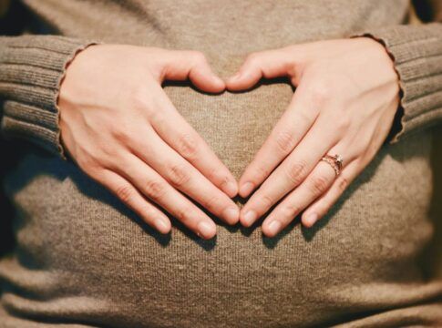 Mãos formando um coração sobre uma barriga de gestante, ambas apoiadas em um suéter de tricô bege; uma das mãos usa um anel delicado.