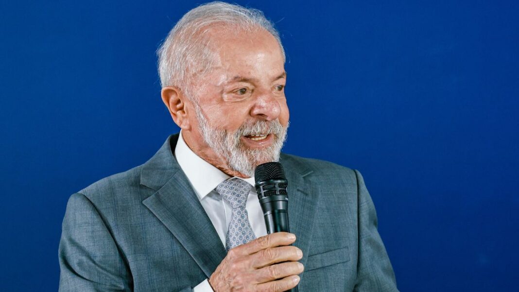 A foto apresenta um plano médio (do peito para cima) de Luiz Inácio Lula da Silva, o atual presidente do Brasil. Ele está posicionado levemente de perfil, voltado para a direita da imagem, enquanto segura um microfone.
