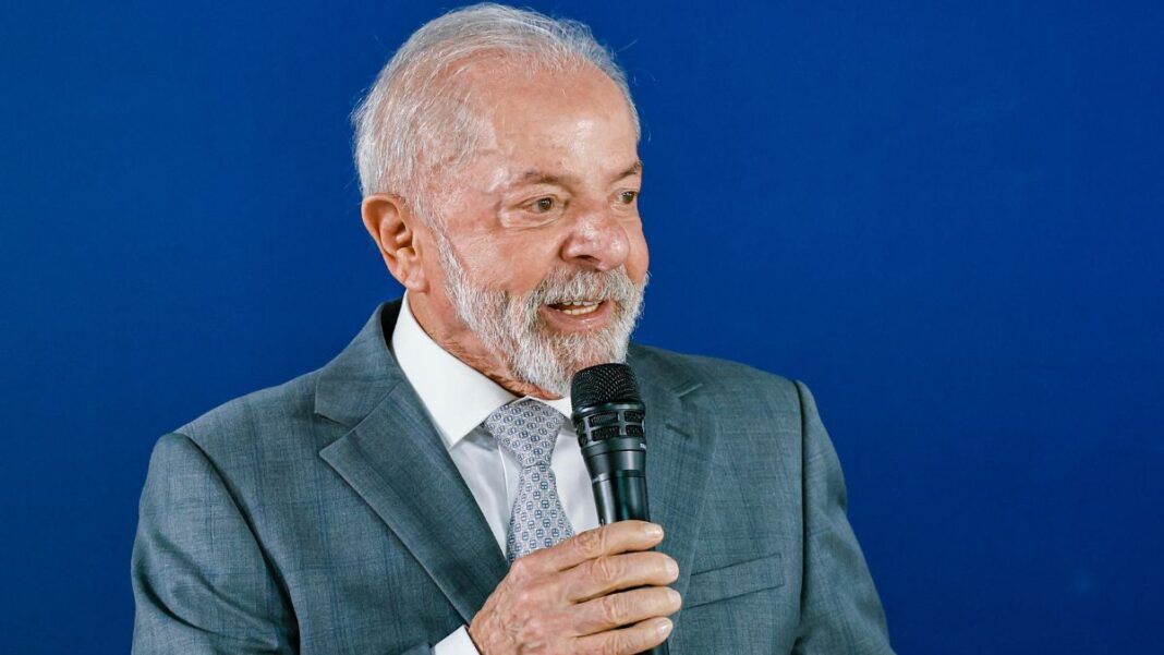 A foto apresenta um plano médio (do peito para cima) de Luiz Inácio Lula da Silva, o atual presidente do Brasil. Ele está posicionado levemente de perfil, voltado para a direita da imagem, enquanto segura um microfone.