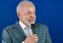 A foto apresenta um plano médio (do peito para cima) de Luiz Inácio Lula da Silva, o atual presidente do Brasil. Ele está posicionado levemente de perfil, voltado para a direita da imagem, enquanto segura um microfone.