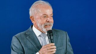 A foto apresenta um plano médio (do peito para cima) de Luiz Inácio Lula da Silva, o atual presidente do Brasil. Ele está posicionado levemente de perfil, voltado para a direita da imagem, enquanto segura um microfone.
