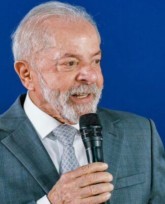 A foto apresenta um plano médio (do peito para cima) de Luiz Inácio Lula da Silva, o atual presidente do Brasil. Ele está posicionado levemente de perfil, voltado para a direita da imagem, enquanto segura um microfone.