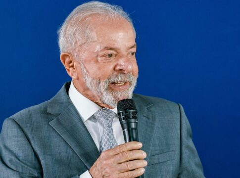 A foto apresenta um plano médio (do peito para cima) de Luiz Inácio Lula da Silva, o atual presidente do Brasil. Ele está posicionado levemente de perfil, voltado para a direita da imagem, enquanto segura um microfone.