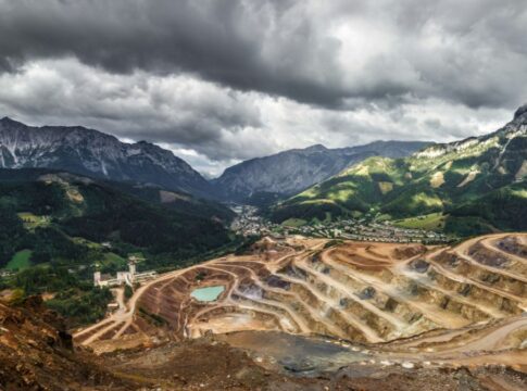 Vista panorâmica de uma grande mina a céu aberto com degraus escavados em tons de marrom e bege; no fundo, há um vale com pequenas construções e, além dele, cadeias de montanhas altas e rochosas sob um céu carregado de nuvens escuras.