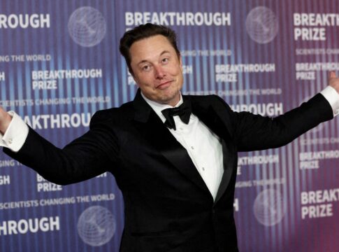 Homem branco vestindo smoking preto com os braços abertos e sorrindo levemente