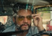 Wagner Moura em cena do filme, dentro de um carro com o vidro sujo, ajustando seu óculos escuro de lente amarelada