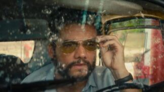 Wagner Moura em cena do filme, dentro de um carro com o vidro sujo, ajustando seu óculos escuro de lente amarelada