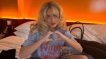 Sabrina Carpenter sentada em uma cama, fazendo um gesto de coração com as mãos. Ela tem cabelo loiro ondulado e usa uma camiseta azul estampada. O ambiente tem iluminação quente alaranjada ao fundo, criando um clima acolhedor. Ao lado dela, há travesseiros, uma bolsa e uma garrafa em um balde.