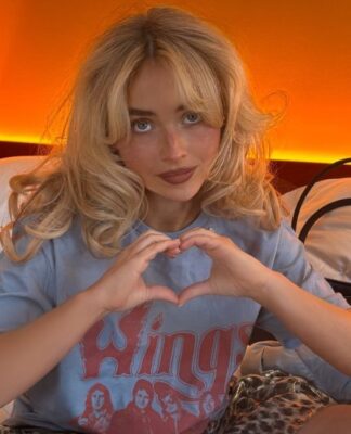 Sabrina Carpenter sentada em uma cama, fazendo um gesto de coração com as mãos. Ela tem cabelo loiro ondulado e usa uma camiseta azul estampada. O ambiente tem iluminação quente alaranjada ao fundo, criando um clima acolhedor. Ao lado dela, há travesseiros, uma bolsa e uma garrafa em um balde.