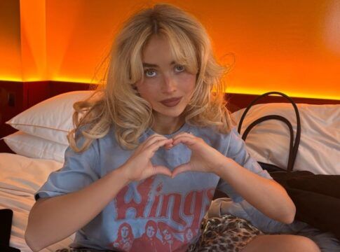 Sabrina Carpenter sentada em uma cama, fazendo um gesto de coração com as mãos. Ela tem cabelo loiro ondulado e usa uma camiseta azul estampada. O ambiente tem iluminação quente alaranjada ao fundo, criando um clima acolhedor. Ao lado dela, há travesseiros, uma bolsa e uma garrafa em um balde.