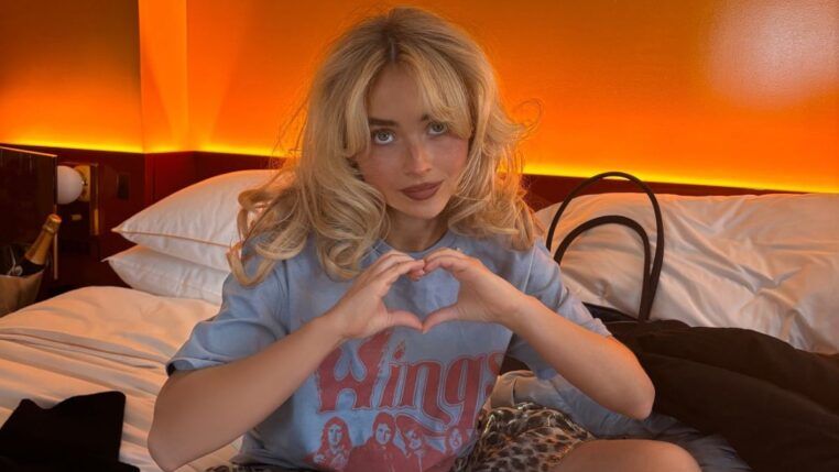Sabrina Carpenter sentada em uma cama, fazendo um gesto de coração com as mãos. Ela tem cabelo loiro ondulado e usa uma camiseta azul estampada. O ambiente tem iluminação quente alaranjada ao fundo, criando um clima acolhedor. Ao lado dela, há travesseiros, uma bolsa e uma garrafa em um balde.
