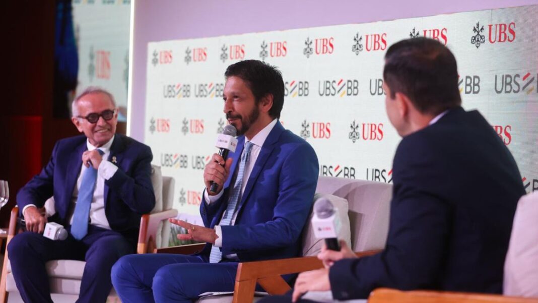 Homens de terno participam de um painel em um evento corporativo, sentados em poltronas e conversando com microfones em mãos. Um painel ao fundo exibe repetidamente os logotipos “UBS” e “UBS BB”. O ambiente é iluminado e formal.