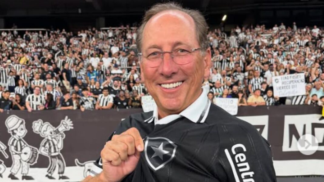 A imagem mostra um homem sorridente usando a camisa preta de um time de futebol, segurando o escudo na altura do peito com orgulho. Ao fundo, há uma grande multidão de torcedores com camisas listradas em preto e branco, criando um clima de festa e apoio no estádio.