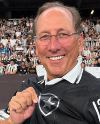 A imagem mostra um homem sorridente usando a camisa preta de um time de futebol, segurando o escudo na altura do peito com orgulho. Ao fundo, há uma grande multidão de torcedores com camisas listradas em preto e branco, criando um clima de festa e apoio no estádio.