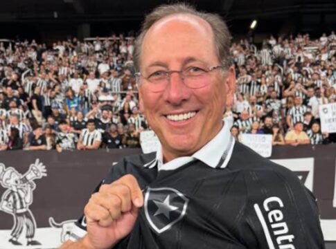 A imagem mostra um homem sorridente usando a camisa preta de um time de futebol, segurando o escudo na altura do peito com orgulho. Ao fundo, há uma grande multidão de torcedores com camisas listradas em preto e branco, criando um clima de festa e apoio no estádio.