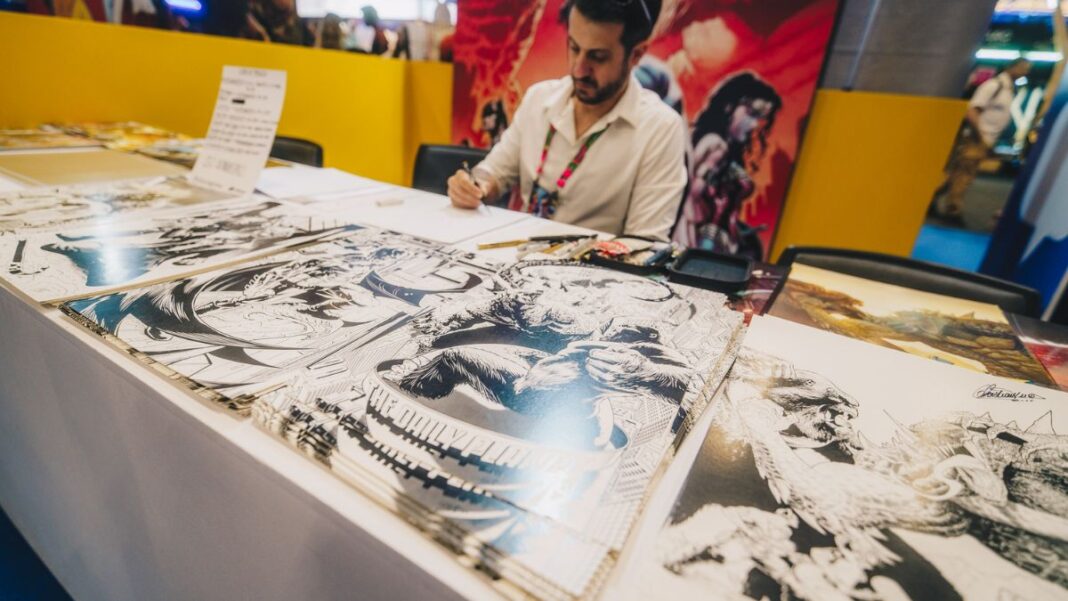 Artista sentado atrás de uma mesa em um evento, desenhando enquanto várias ilustrações em preto e branco de estilo quadrinhos estão expostas à sua frente; ao fundo, painéis coloridos com arte de personagens.