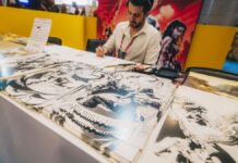 Artista sentado atrás de uma mesa em um evento, desenhando enquanto várias ilustrações em preto e branco de estilo quadrinhos estão expostas à sua frente; ao fundo, painéis coloridos com arte de personagens.