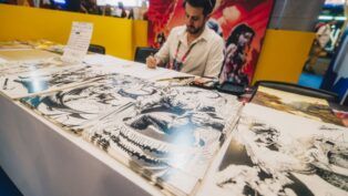 Artista sentado atrás de uma mesa em um evento, desenhando enquanto várias ilustrações em preto e branco de estilo quadrinhos estão expostas à sua frente; ao fundo, painéis coloridos com arte de personagens.