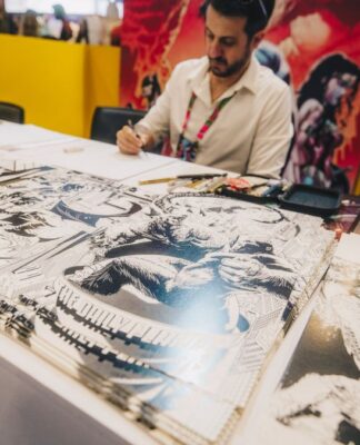 Artista sentado atrás de uma mesa em um evento, desenhando enquanto várias ilustrações em preto e branco de estilo quadrinhos estão expostas à sua frente; ao fundo, painéis coloridos com arte de personagens.
