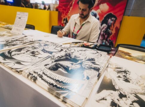 Artista sentado atrás de uma mesa em um evento, desenhando enquanto várias ilustrações em preto e branco de estilo quadrinhos estão expostas à sua frente; ao fundo, painéis coloridos com arte de personagens.