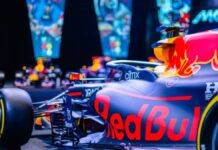 Carro de Fórmula 1 da Red Bull em destaque, com pintura azul e detalhes em vermelho e amarelo, exibido em ambiente iluminado com telas coloridas ao fundo mostrando imagens relacionadas à equipe.