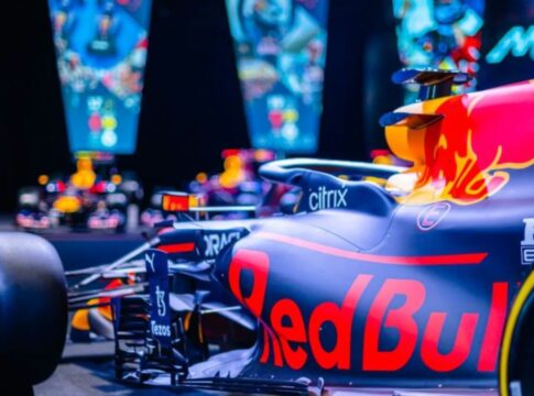 Carro de Fórmula 1 da Red Bull em destaque, com pintura azul e detalhes em vermelho e amarelo, exibido em ambiente iluminado com telas coloridas ao fundo mostrando imagens relacionadas à equipe.